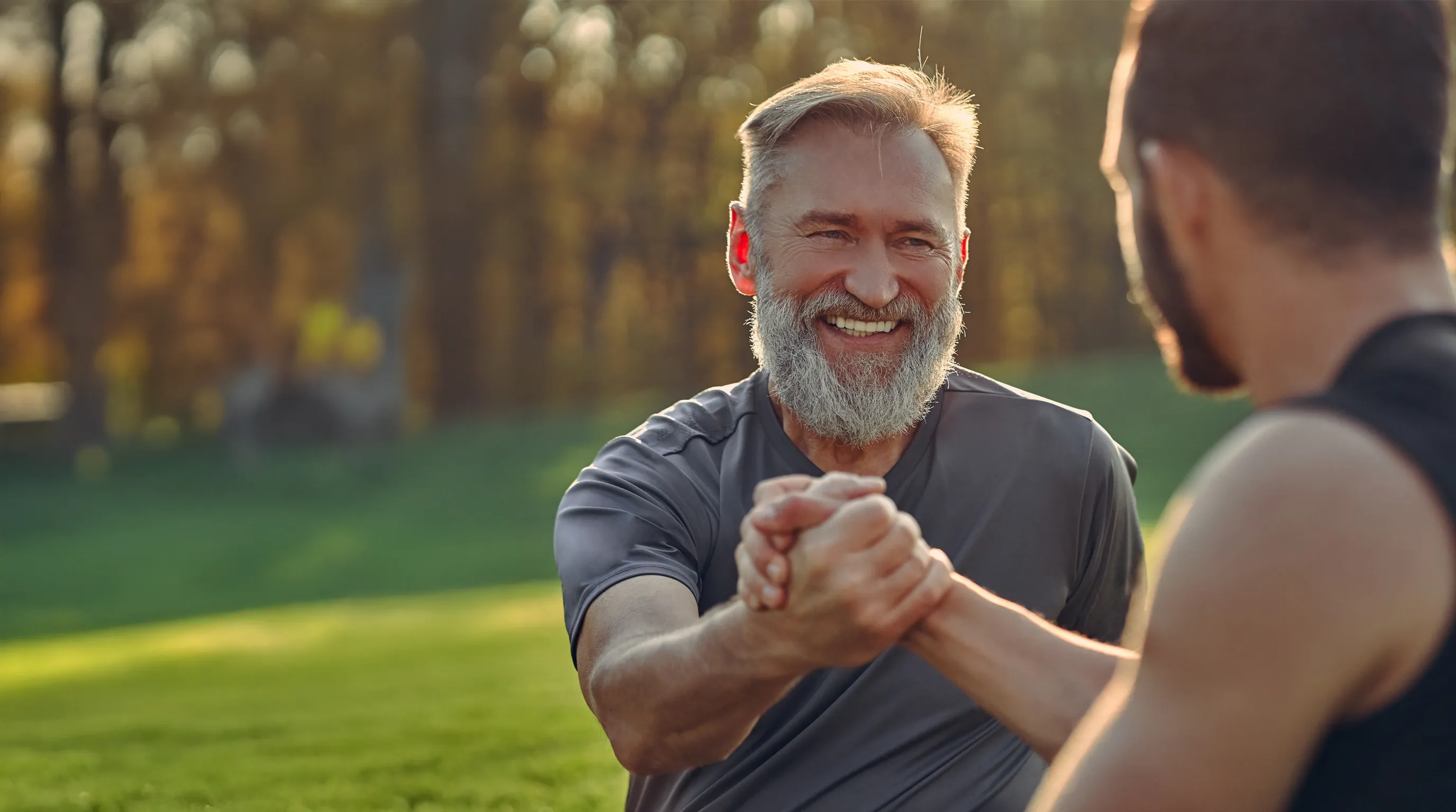 An older man laughing warmly while clasping a friend's hand outdoors at golden hour — connection, vitality, hope.