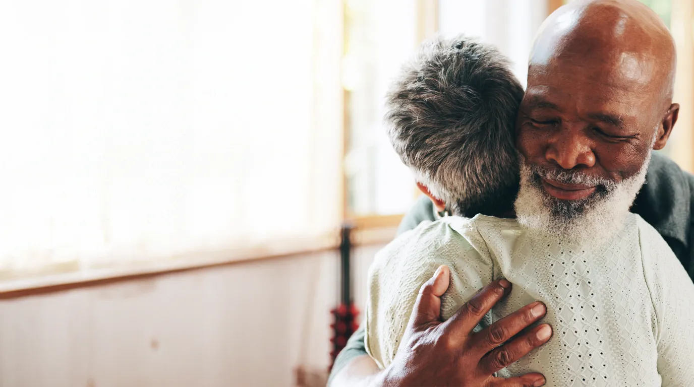 An older couple sharing a quiet, close embrace by a sunlit window — natural light, warm interior.